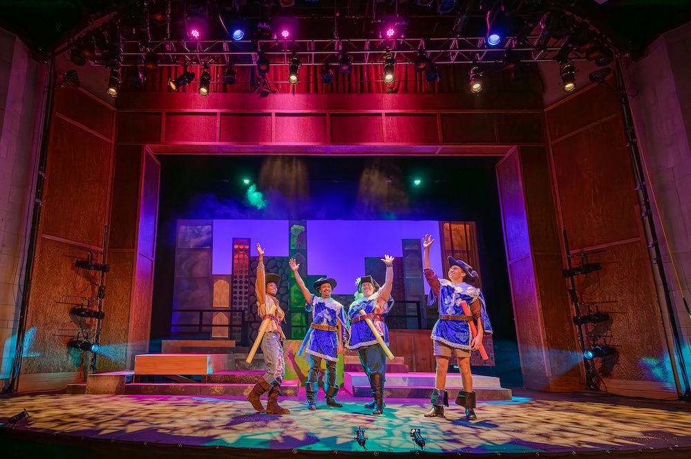 Four students dressed as Muskateers raise their hands on stage